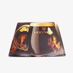 May include: A vintage vinyl record album cover featuring a large glass of Guinness beer with a group of people in the background. The album cover is black with a gold Guinness logo.