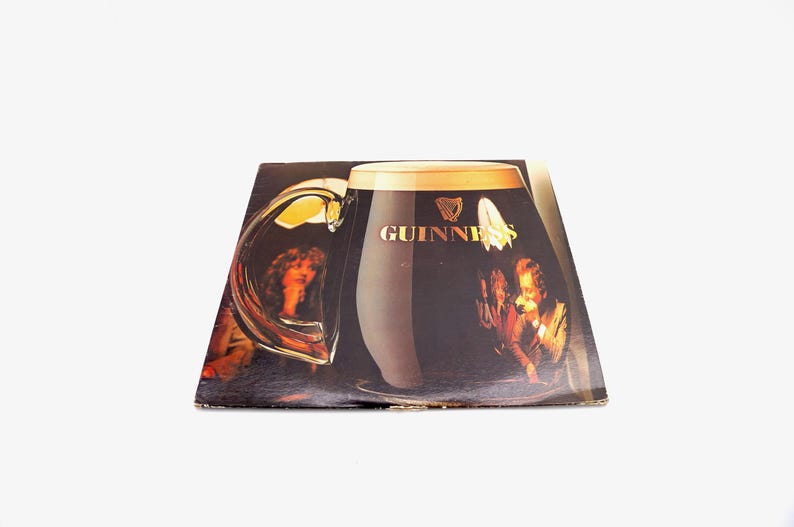 May include: A vintage vinyl record album cover featuring a large glass of Guinness beer with a group of people in the background. The album cover is black with a gold Guinness logo.