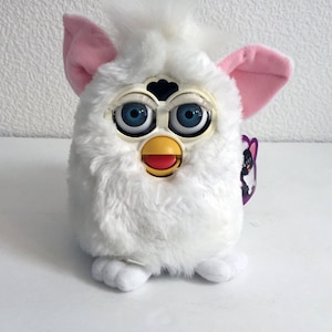 May include: White furry interactive toy with blue eyes, a yellow beak, and a red mouth. The toy has pink ears and a purple tag with a black and white image.