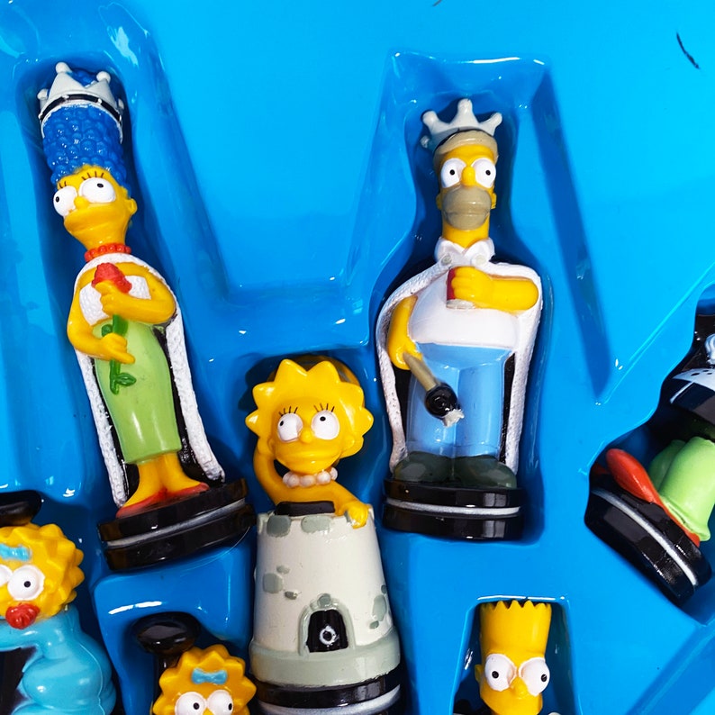 May include: A set of Simpsons character figurines, including Homer, Marge, Lisa, and Bart, in a blue plastic tray. The figurines are yellow and have their signature hairstyles.