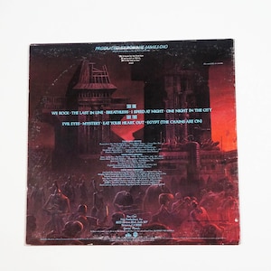 May include: Back cover of a vinyl record album with a dark, red, and orange illustration of a city with tall buildings and a crowd of people in the foreground. The album is titled "Produced by Ronnie James Dio" and lists the songs "We Rock", "The Last in Line", "BreathLess", "I Speed at Night", "One Night in the City", "Evil Eyes", "Mystery", "Eat Your Heart Out", and "Egypt (The Chains Are On)".