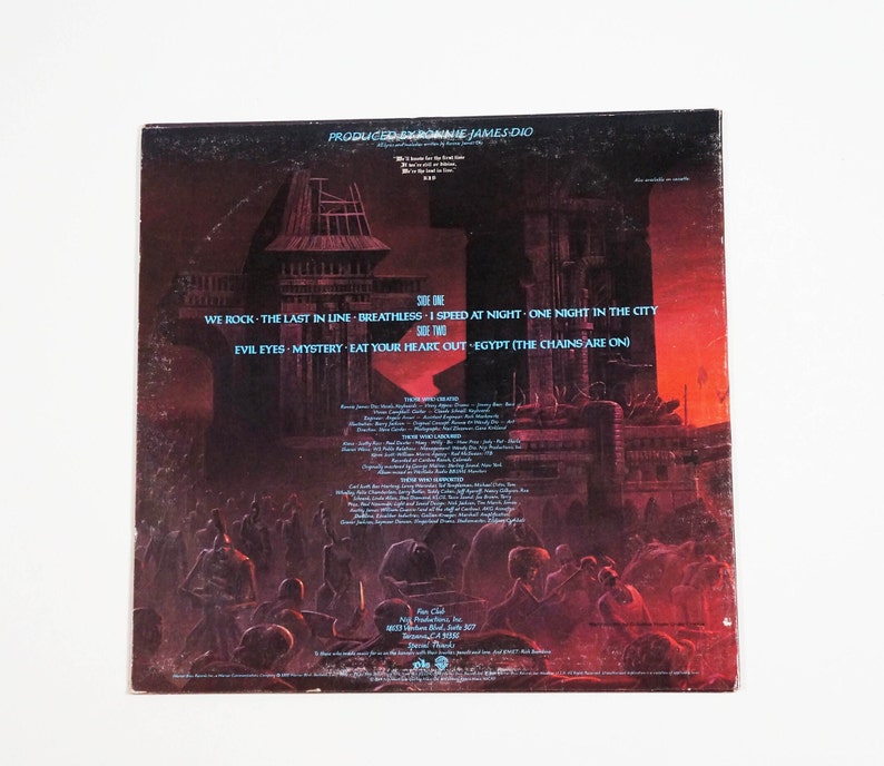 May include: Back cover of a vinyl record album with a dark, red, and orange illustration of a city with tall buildings and a crowd of people in the foreground. The album is titled "Produced by Ronnie James Dio" and lists the songs "We Rock", "The Last in Line", "BreathLess", "I Speed at Night", "One Night in the City", "Evil Eyes", "Mystery", "Eat Your Heart Out", and "Egypt (The Chains Are On)".