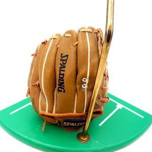 K&ouml;nnte beinhalten: Ein brauner Spalding-Baseballhandschuh aus Leder mit wei&szlig;en N&auml;hten und orangefarbenen Akzenten. Der Handschuh wird auf einer gr&uuml;nen, gebogenen Basis mit wei&szlig;en Linien und von einem goldfarbenen Metallst&auml;nder getragen.