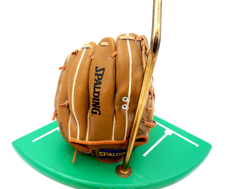 K&ouml;nnte beinhalten: Ein brauner Spalding-Baseballhandschuh aus Leder mit wei&szlig;en N&auml;hten und orangefarbenen Akzenten. Der Handschuh wird auf einer gr&uuml;nen, gebogenen Basis mit wei&szlig;en Linien und von einem goldfarbenen Metallst&auml;nder getragen.