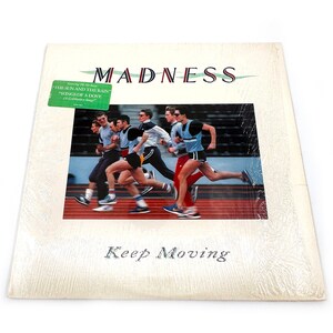 May include: A vintage vinyl record album cover featuring a group of men running on a red track. The album cover is white with the band name "MADNESS" in large, bold, red letters. The album title "Keep Moving" is printed in black below the image. The album features the songs "The Sun and the Rain" and "Wings of a Dove".