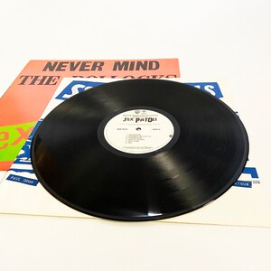 May include: A black vinyl record with a white label that says "Never Mind The Bollocks" and "Sex Pistols". The record is on top of a white record sleeve with the text "Never Mind The Bollocks" and "Sex Pistols" printed on it.