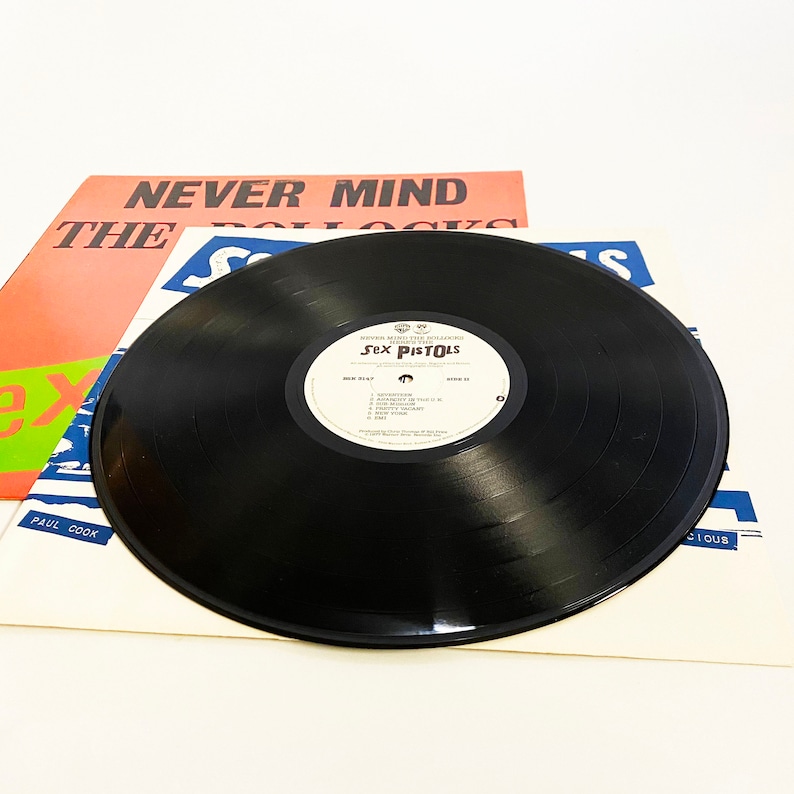 May include: A black vinyl record with a white label that says "Never Mind The Bollocks" and "Sex Pistols". The record is on top of a white record sleeve with the text "Never Mind The Bollocks" and "Sex Pistols" printed on it.