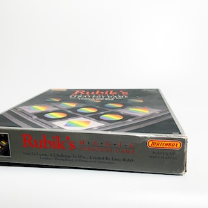 May include: A silver game box with a rainbow design. The box is for Rubik's Magic Strategy Game, created by Erno Rubik. The box says "Easy to Learn, A Challenge to Win..." and "Ages 8 & Up, For 2 Players".