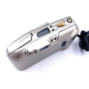 May include: A silver point-and-shoot film camera with a black wrist strap. The camera has a digital display and several buttons, including a mode set button. The text "PANORAMA" and "QUARTZDATE MODE SET" are visible on the camera.