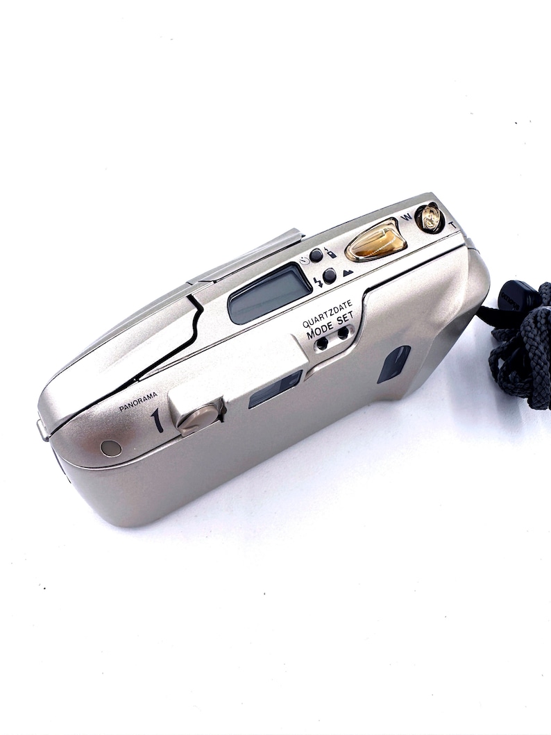 May include: A silver point-and-shoot film camera with a black wrist strap. The camera has a digital display and several buttons, including a mode set button. The text "PANORAMA" and "QUARTZDATE MODE SET" are visible on the camera.