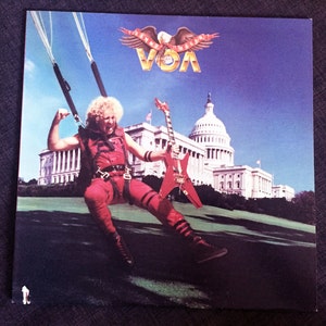 May include: A red, white, and blue album cover featuring a man in a red jumpsuit holding a red electric guitar. The man is suspended in the air by a parachute. The Capitol Building is in the background. The album cover is titled "Sammy Hagar - VOA".