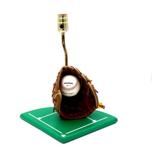 K&ouml;nnte beinhalten: Baseball-Themenlampe mit einem braunen Baseballhandschuh aus Leder, der einen Baseball h&auml;lt. Der Baseball tr&auml;gt den Text "CHAMPRO". Die Lampe ist auf einer gr&uuml;nen, rautenf&ouml;rmigen Basis montiert.