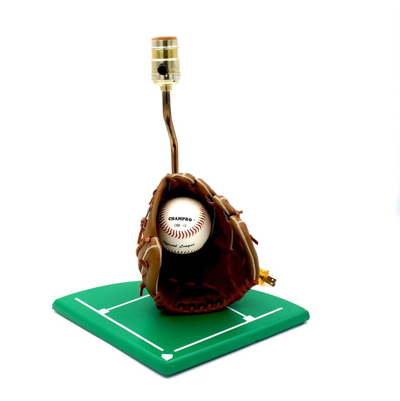 K&ouml;nnte beinhalten: Baseball-Themenlampe mit einem braunen Baseballhandschuh aus Leder, der einen Baseball h&auml;lt. Der Baseball tr&auml;gt den Text "CHAMPRO". Die Lampe ist auf einer gr&uuml;nen, rautenf&ouml;rmigen Basis montiert.