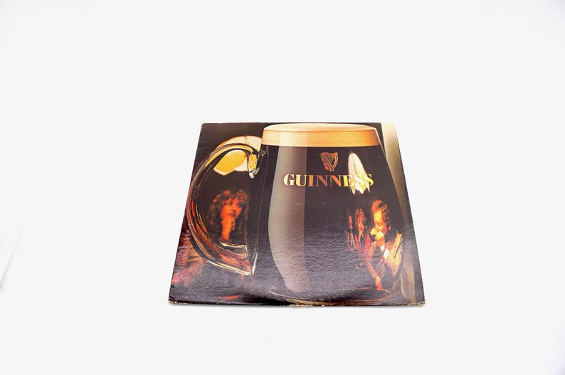 May include: A vintage vinyl record album cover featuring a large glass of Guinness beer with a woman's face reflected in the glass. The album cover is brown and black with the Guinness logo and text.