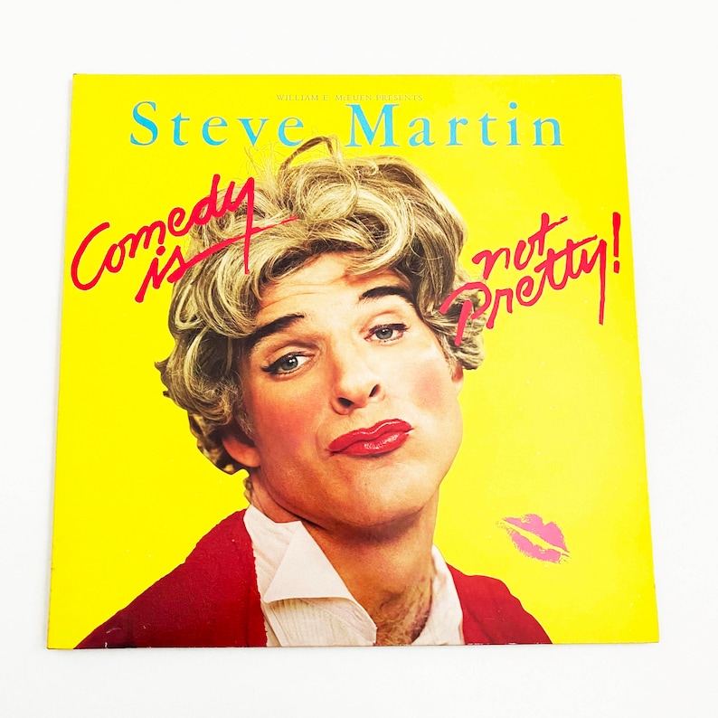 May include: A yellow album cover with a man wearing a red jacket and a blonde wig. The text on the cover reads "Steve Martin Comedy is not Pretty!"