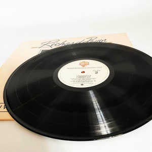 Puede incluir: Disco de vinilo negro con una etiqueta blanca. La etiqueta tiene el texto "Richard Pryor Live On The Sunset Strip" y "Side 2".
