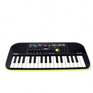 May include: Black Casio SA-46 keyboard with white keys and a yellow accent. The keyboard has a digital display and various function buttons. The model name is visible on the keyboard.