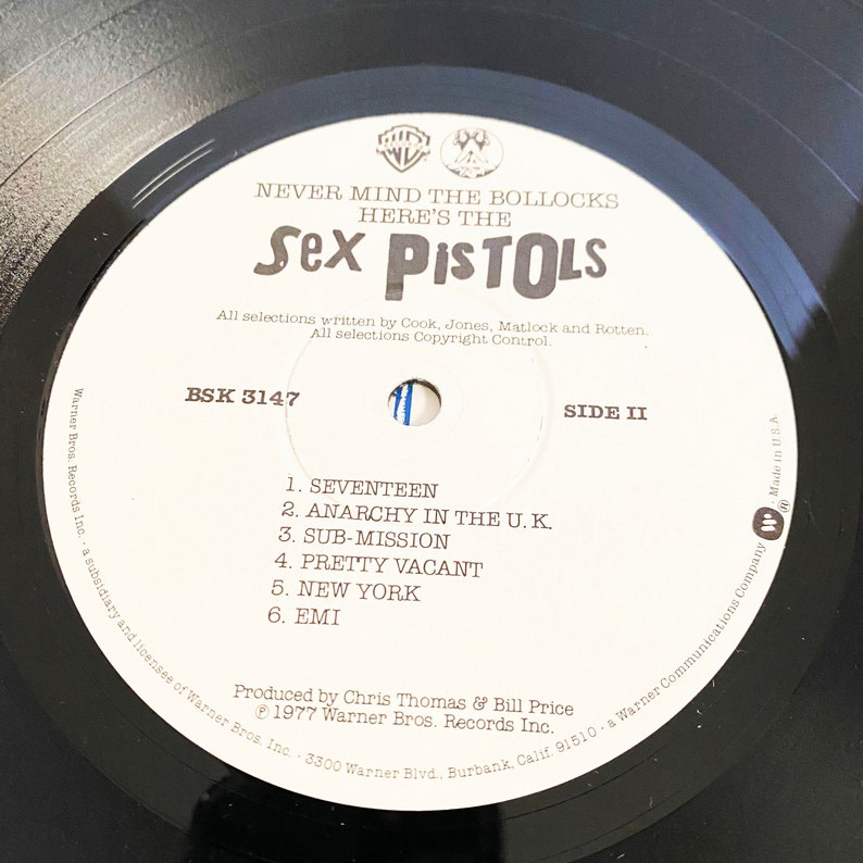 May include: Black and white vinyl record with a white label. The label has the text "Never Mind The Bollocks Here's The Sex Pistols" and the track list for side two of the record. The track list includes "Seventeen", "Anarchy In The U.K.", "Sub-Mission", "Pretty Vacant", "New York", and "EMI".