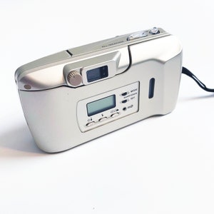 May include: A silver compact camera with a built-in flash and a digital display. The camera has a date and time setting and a mode dial. The camera is labeled "All-Weather".