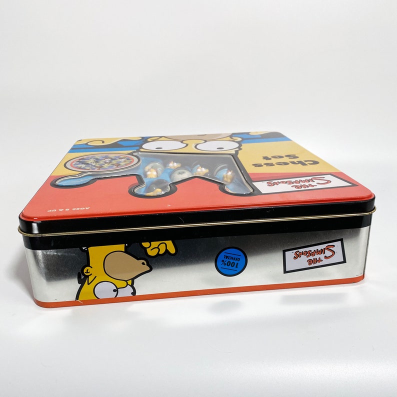 May include: A yellow, red, and blue tin box with a picture of Homer Simpson on the front. The box says "The Simpsons Chess Set" and "100% Official".