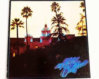 May include: A vintage vinyl record album cover featuring a nighttime scene of a building with palm trees and the text "Hotel California" in blue and purple lettering.