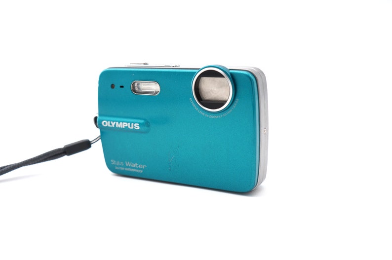 May include: A teal blue Olympus Stylus Water digital camera with a silver lens ring and a black wrist strap. The camera has the text "Olympus" and "Stylus Water" printed on the front.