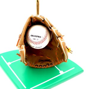 K&ouml;nnte beinhalten: Ein Baseballhandschuh mit einem Baseball darin, der auf einem gr&uuml;nen Quadrat mit wei&szlig;en Linien ruht. Der Baseball ist wei&szlig; mit roten N&auml;hten und dem Text "CHAMPRO". Der Handschuh ist braun mit orangefarbenen N&auml;hten. Die Basis ist 10 cm x 10 cm gro&szlig;.