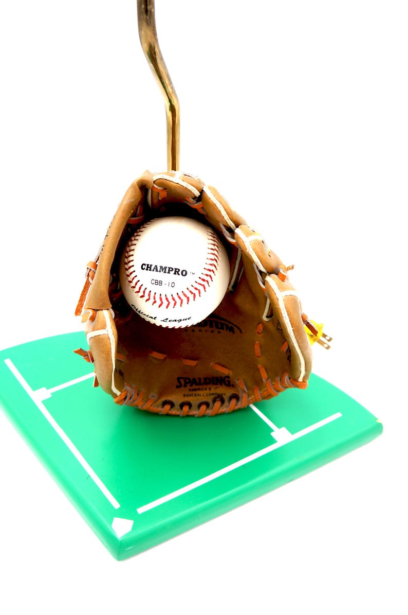 K&ouml;nnte beinhalten: Ein Baseballhandschuh mit einem Baseball darin, der auf einem gr&uuml;nen Quadrat mit wei&szlig;en Linien ruht. Der Baseball ist wei&szlig; mit roten N&auml;hten und dem Text "CHAMPRO". Der Handschuh ist braun mit orangefarbenen N&auml;hten. Die Basis ist 10 cm x 10 cm gro&szlig;.