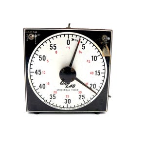 May include: A vintage black and white Universal Timer by Lab, with a circular dial and a black frame. The dial features numbers in red and black, with a central clock hand. The timer has an on/off switch and outlet plug.