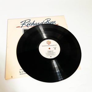 Puede incluir: Un disco de vinilo negro con una etiqueta blanca que dice "Richard Pryor Live on the Sunset Strip" y "Warner Bros. Records" en letras rojas y azules. El disco est&aacute; sobre una superficie blanca con un papel blanco con el texto "Richard Pryor" en negro.