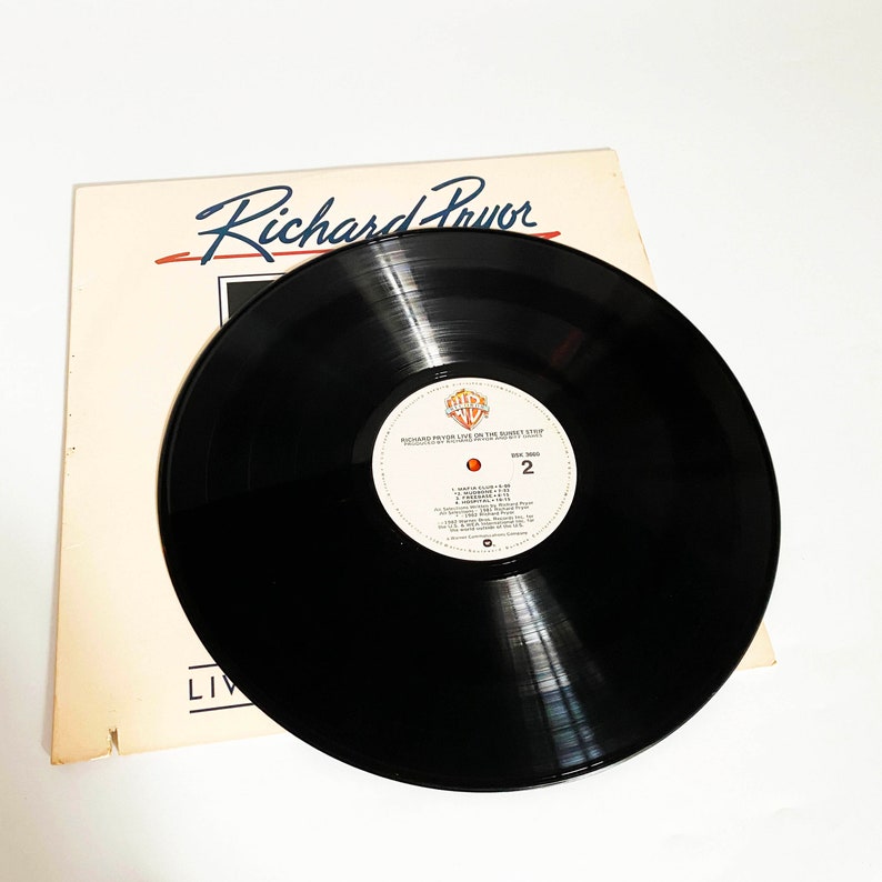 Puede incluir: Un disco de vinilo negro con una etiqueta blanca que dice "Richard Pryor Live on the Sunset Strip" y "Warner Bros. Records" en letras rojas y azules. El disco est&aacute; sobre una superficie blanca con un papel blanco con el texto "Richard Pryor" en negro.