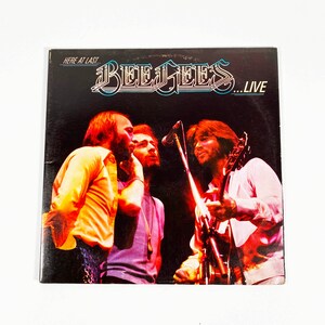 May include: A vintage vinyl record album cover featuring a live performance by the band, The Bee Gees. The cover features a black background with the band members in a concert setting. The album title, "Here At Last... Bee Gees... Live", is printed in white and blue lettering.