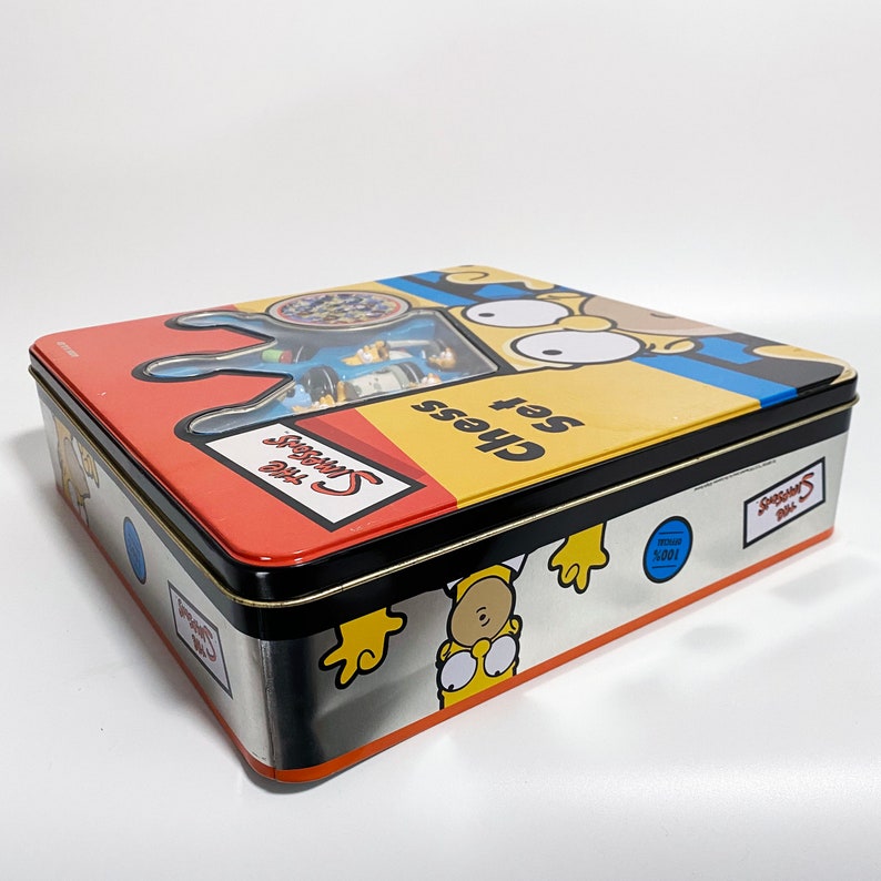 May include: A yellow, blue, and red metal tin box with the Simpsons characters on it. The box says "The Simpsons Chess Set" on the front.