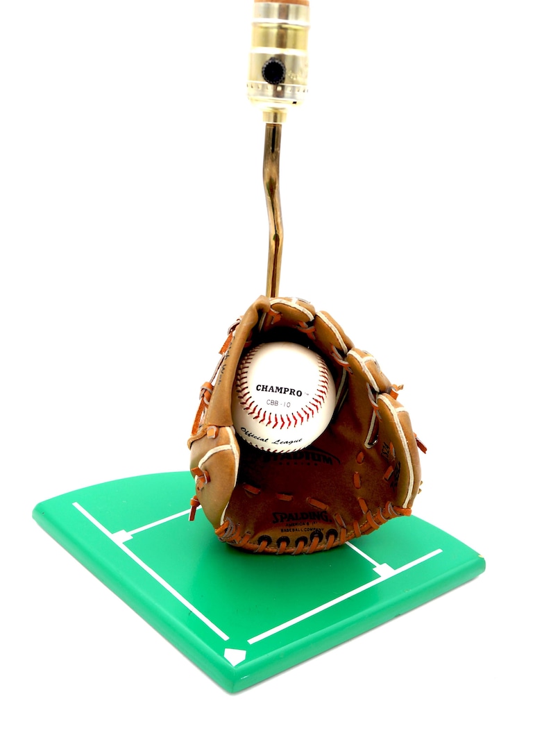 K&ouml;nnte beinhalten: Baseball-Themenlampe. Ein brauner Baseballhandschuh aus Leder h&auml;lt einen Baseball mit dem Text "CHAMPRO CBB 10 Official League". Die Lampe ist auf einer gr&uuml;nen Basis in Form eines Baseballfelds montiert.