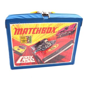 May include: Blue Matchbox Carry Case with a red, yellow, and black race car on the front. The case has a yellow handle and a silver latch. The text on the front reads "Matchbox" and "Carry Case".