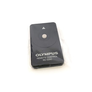 May include: Black Olympus RC-300C remote control in a clear plastic package. The remote has a circular button and an arrow symbol. The text "OLYMPUS REMOTE CONTROL RC-300C" is printed on the front.