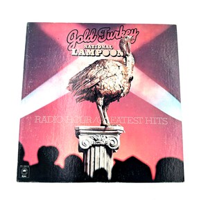 May include: Album cover for "Gold Turkey National Lampoon Radio Hour/Greatest Hits." The cover features a golden turkey statue on a pedestal against a red and pink background. The album title is in a stylized font, with the text "Radio Hour/Greatest Hits" below.