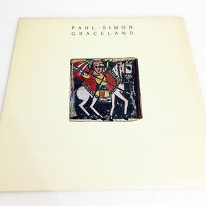 May include: A white vinyl record album cover with the title "PAUL SIMON GRACELAND" printed in black text. The cover features a colorful illustration of a person riding a white horse.