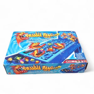 May include: A boxed board game titled "Piranha Panic". The box features a blue background with cartoon piranhas and fish. The game's title and tagline, "Race to escape the feeding frenzy!", are displayed on the box.
