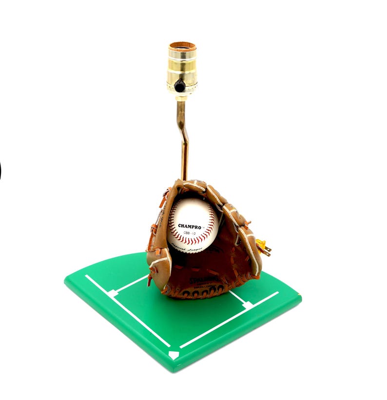 K&ouml;nnte beinhalten: Baseball-Themenlampe mit einem braunen Baseballhandschuh aus Leder, der einen Baseball h&auml;lt. Die Lampe hat einen messingfarbenen Hals und einen zylindrischen Lampenhalter. Der Sockel ist ein gr&uuml;nes, rautenf&ouml;rmiges Feld.