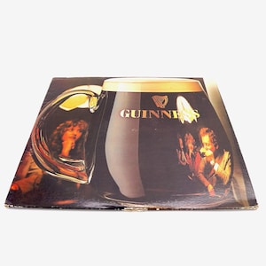 May include: A vintage vinyl record album cover featuring a large glass of Guinness beer with the Guinness logo and a harp. The glass is partially filled with dark brown liquid and has a white foam head. The background shows a group of people in a dimly lit setting.