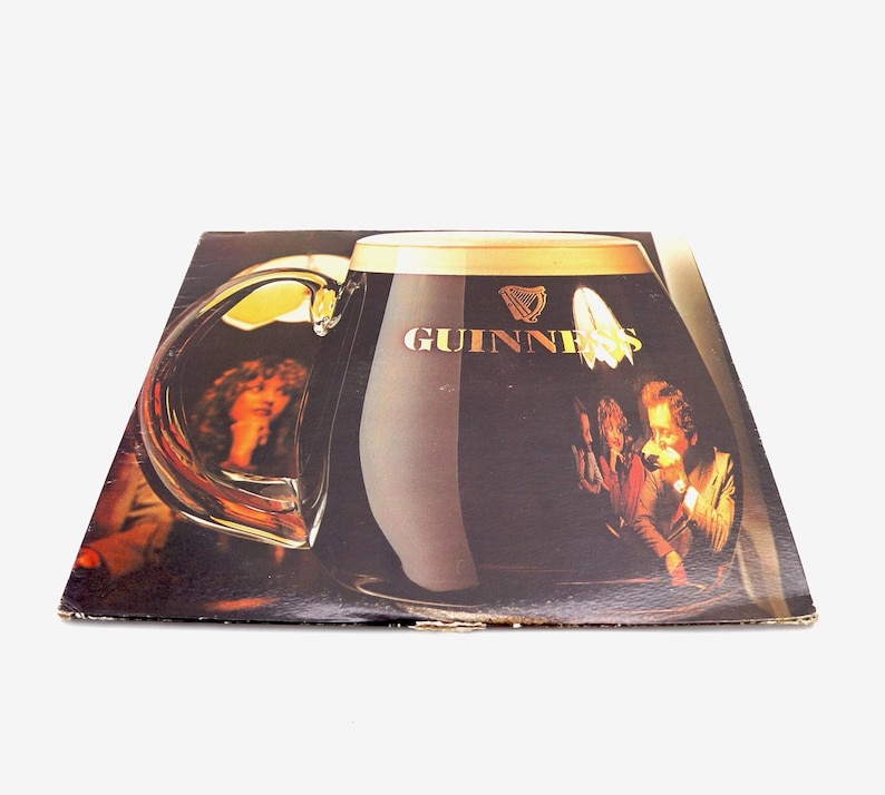 May include: A vintage vinyl record album cover featuring a large glass of Guinness beer with the Guinness logo and a harp. The glass is partially filled with dark brown liquid and has a white foam head. The background shows a group of people in a dimly lit setting.