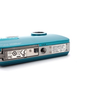 May include: A teal blue Olympus digital camera with the text "Stylus Water" and "Olympus Imaging Corp. Digital Camera" printed on the back. The camera has a silver trim and a black strap.