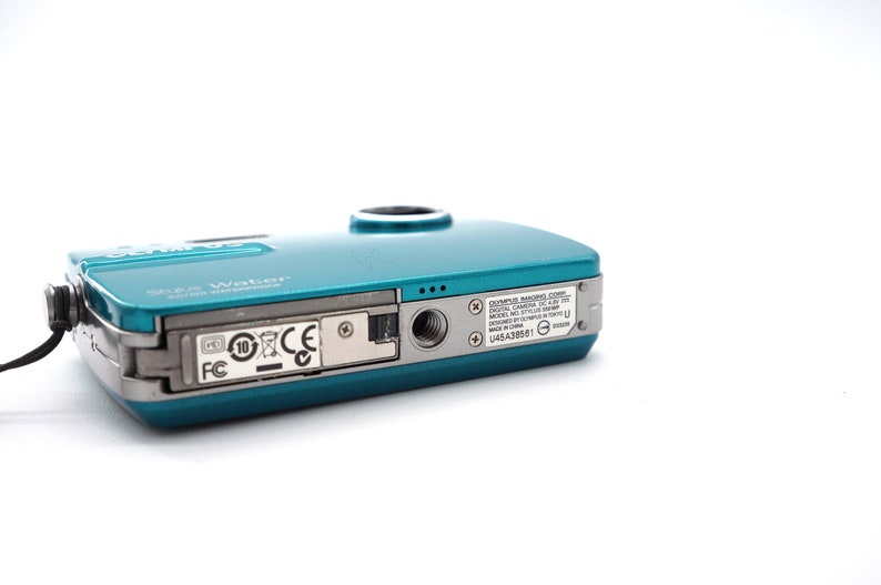 May include: A teal blue Olympus digital camera with the text "Stylus Water" and "Olympus Imaging Corp. Digital Camera" printed on the back. The camera has a silver trim and a black strap.