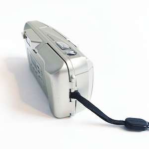 May include: A silver point-and-shoot camera with a black strap. The camera has a built-in flash and a zoom lens. The camera is made in China.