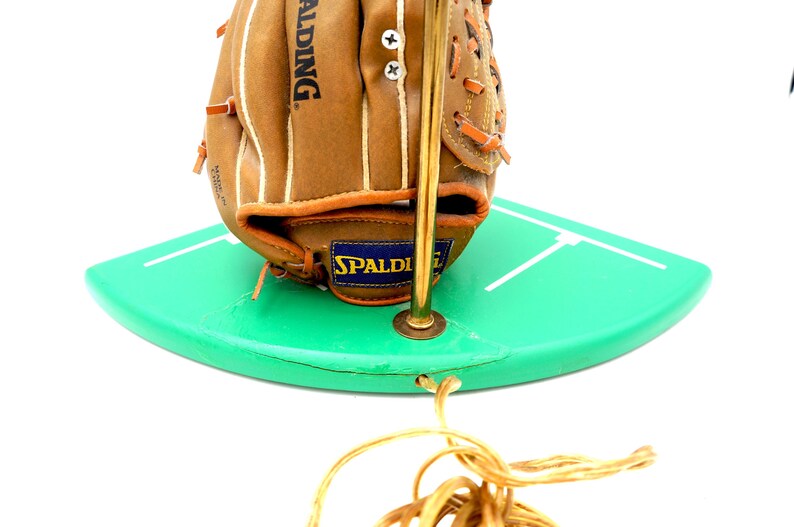 K&ouml;nnte beinhalten: Ein Baseballhandschuh mit dem Spalding-Logo liegt auf einer gr&uuml;nen, gebogenen Oberfl&auml;che mit wei&szlig;en Linien. Der Handschuh ist braun mit wei&szlig;en N&auml;hten, und eine goldfarbene Metallstange ist befestigt. Unten befindet sich eine goldfarbene Schnur.