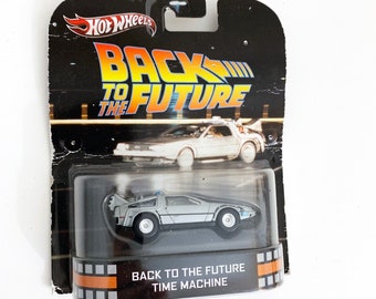 May include: A silver Hot Wheels die-cast toy car of the DeLorean time machine from the movie Back to the Future. The car is in a plastic display case with a black and orange background. The packaging features the movie title "Back to the Future" and the words "Time Machine".