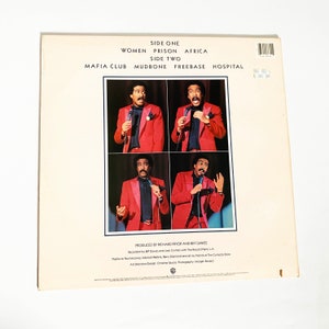 Puede incluir: Una portada de &aacute;lbum de vinilo vintage con el comediante Richard Pryor. La portada presenta cuatro fotos de Pryor con un traje rojo y corbata, actuando en el escenario. El t&iacute;tulo del &aacute;lbum es "Women Prison Africa" y "Mafia Club Mudbone Freebase Hospital".