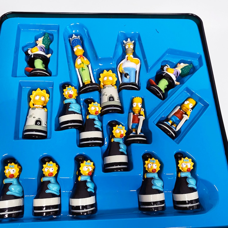 May include: A blue and yellow Simpsons chess set with all the pieces in place on a blue board. The pieces are made of plastic and feature the characters from the Simpsons television show.