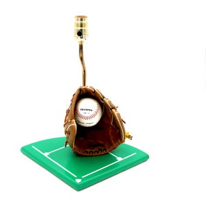 K&ouml;nnte beinhalten: Eine Baseball-Lampe mit einem Baseballhandschuh, der einen Baseball h&auml;lt. Der Baseball tr&auml;gt den Text "CHAMPRO Official League". Die Lampe steht auf einer gr&uuml;nen, rautenf&ouml;rmigen Basis.
